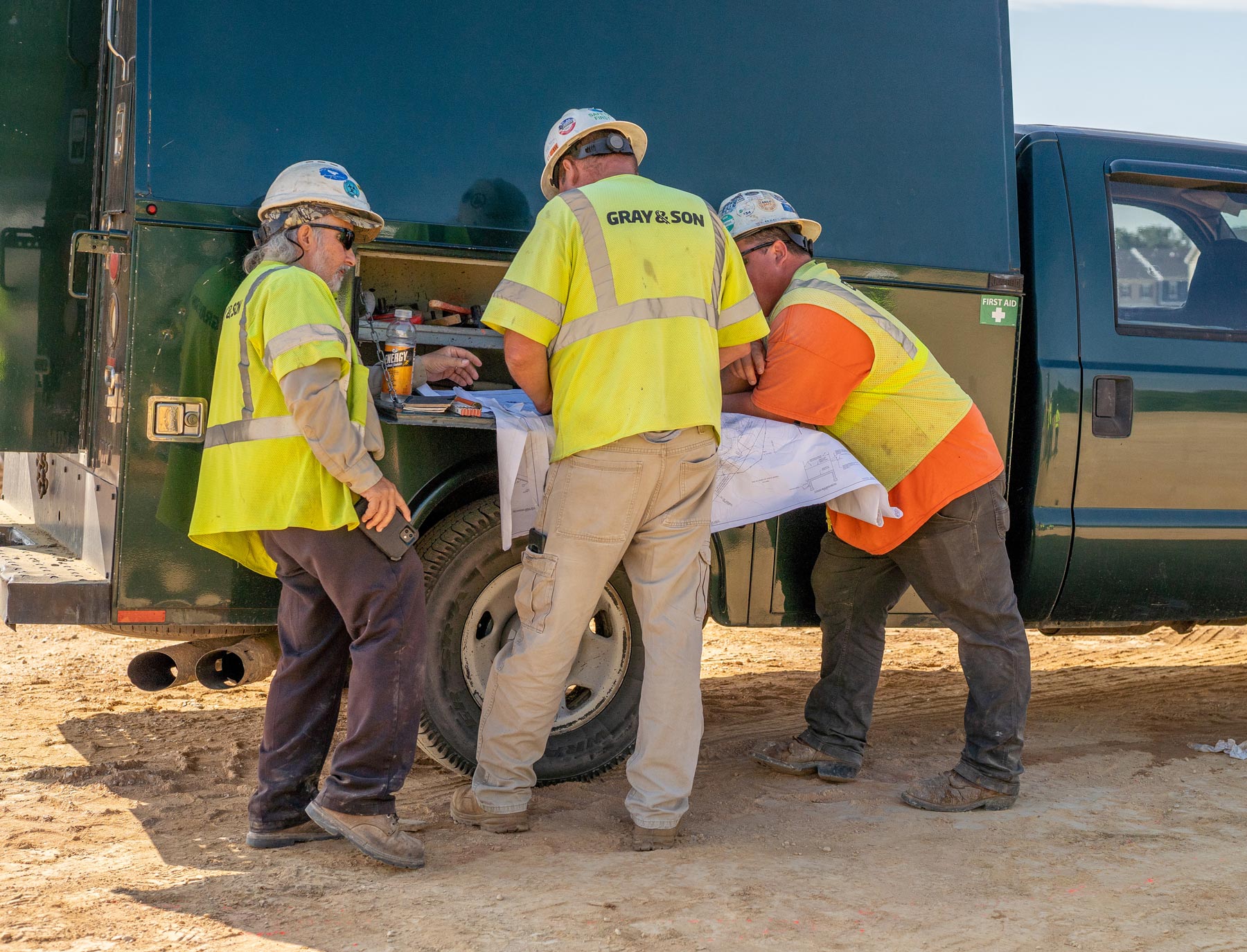 Three males on a construction site conversing by their truck about a blueprint working for a company named Gray & Son a leading site and development contractor in Maryland.