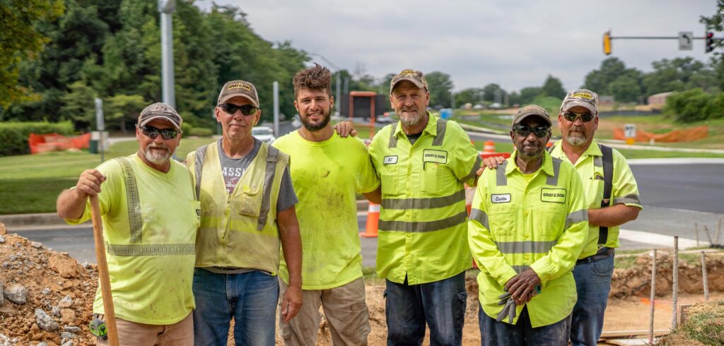 Five male construction workers wearing yellow construction vests smiling for the camera on a construction site, they work for Gray & Son a leading site and development contractor in Maryland.