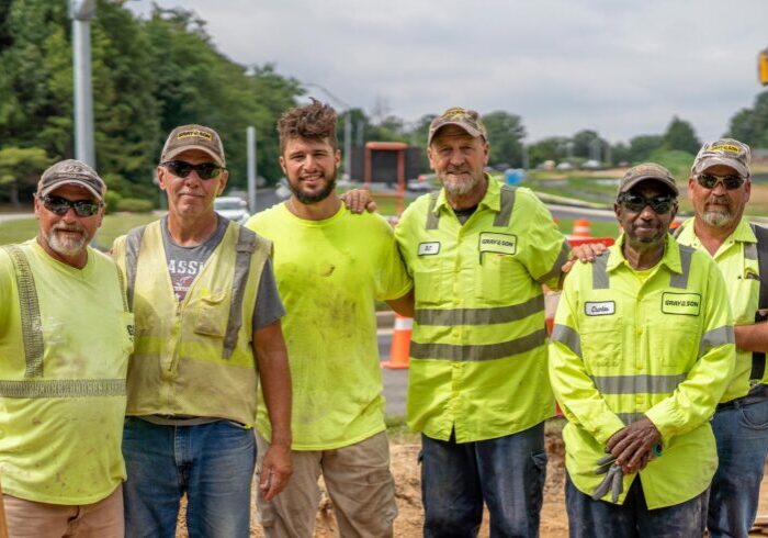 Five male construction workers wearing yellow construction vests smiling for the camera on a construction site, they work for Gray & Son a leading site and development contractor in Maryland.